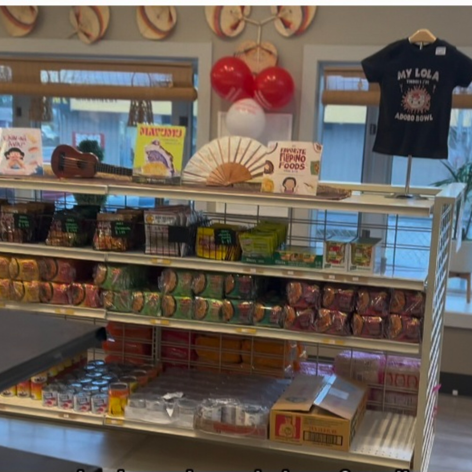 A retail shelf at Kuya's Islander Cuisine filled with Filipino food products, books, and a ukulele.