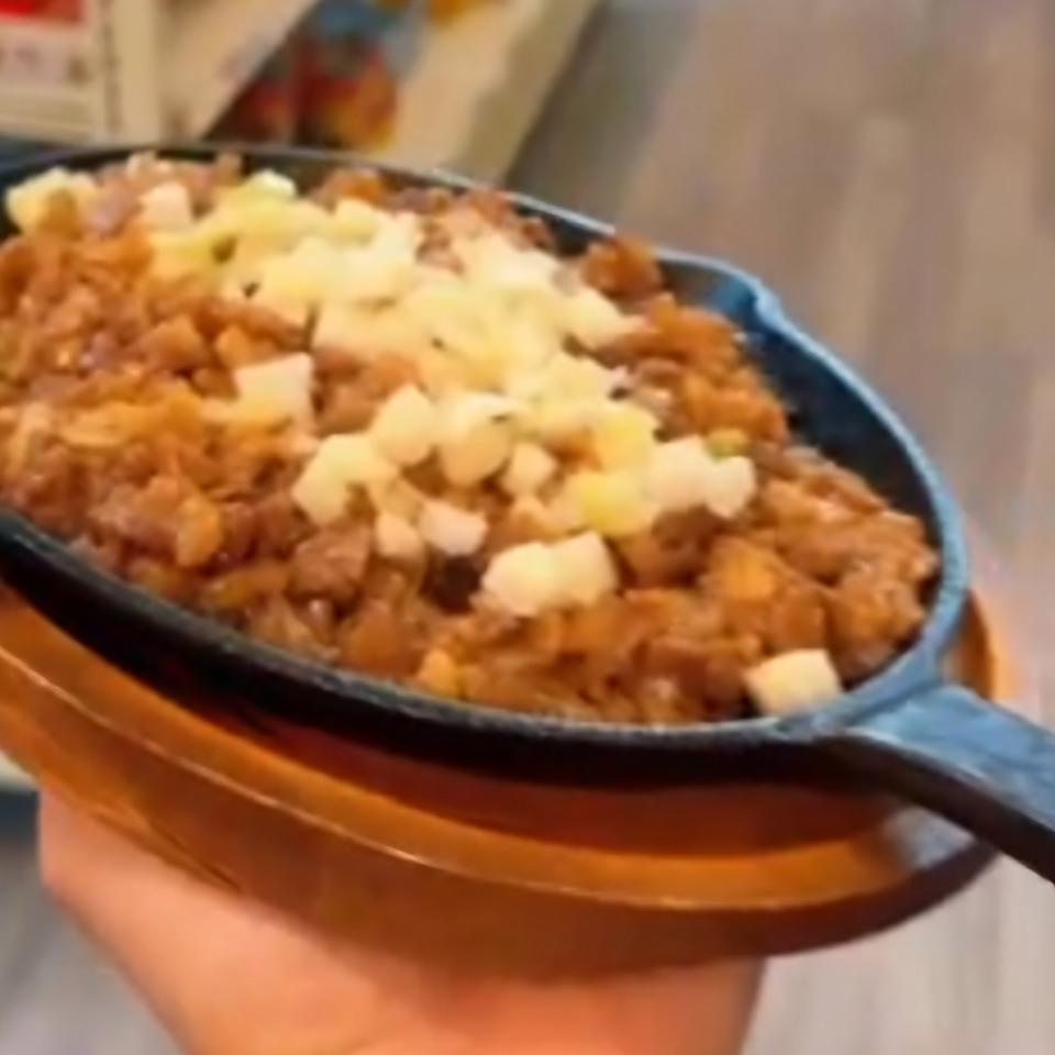 A sizzling plate of Filipino sisig, topped with diced cheese, held by a hand.