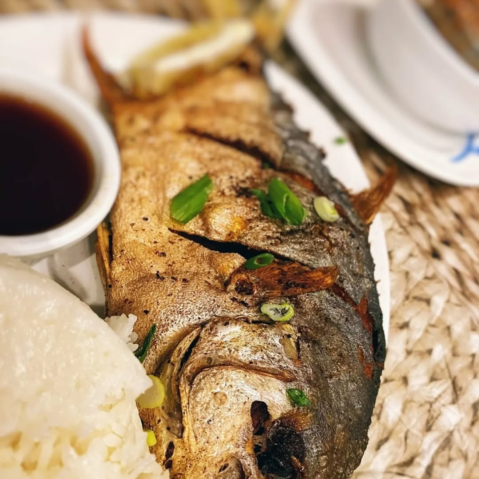 Fried whole fish with rice, a side of sauce, and lemon wedges on a woven placemat.